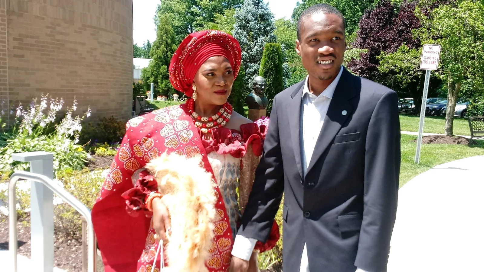 Antonia Akwule, founder of Gregory & Friends, in traditional red Nigerian attire and gele, standing beside her son Gregory in a dark suit on a sunny afternoon